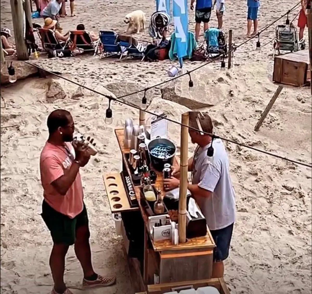 An exterior beach bar scene showcasing two men engaged in conversation at the bar, drinks and mixers set up on the counter, and several people relaxing in beach chairs nearby. The environment is open and festive, with the beach and ocean visible in the background.