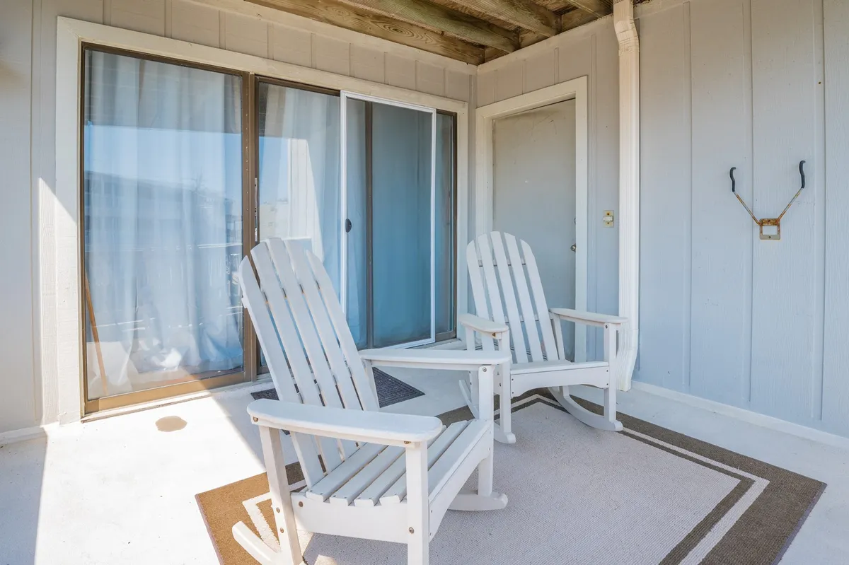 The scene shows two white Adirondack chairs set on a porch outside a sliding glass door, with a neutral beige rug underfoot. The chairs are arranged for relaxed conversation or quiet enjoyment, suggesting a perfect spot to sip coffee as the sun rises. The atmosphere is peaceful, bright, and inviting.