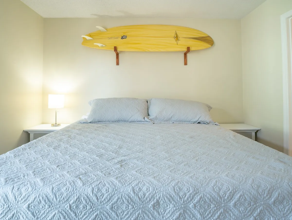 A tranquil corner bedroom featuring a twin bed with a light blue quilt, surfboard wall decor, and a bedside lamp. The setup is simple but stylish, evoking restful beach nights or providing a flexible sleeping arrangement for guests or children.