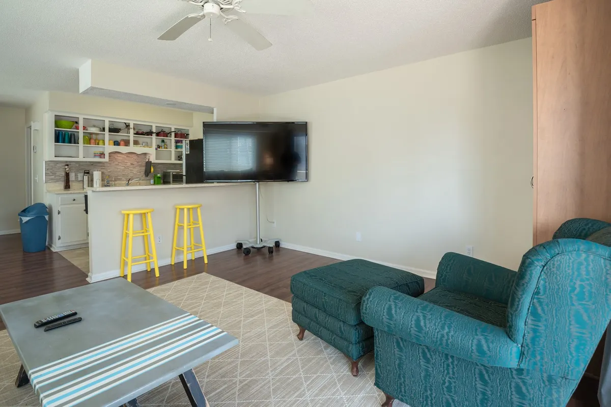 This image presents an inviting open-plan living area featuring a comfortable sofa, rustic wood coffee table, smart TV setup, and a glimpse of the adjacent kitchen with bar stools. Light fills the room, suggesting a casual, sociable hub perfect for movie nights, work, or relaxing afternoons.