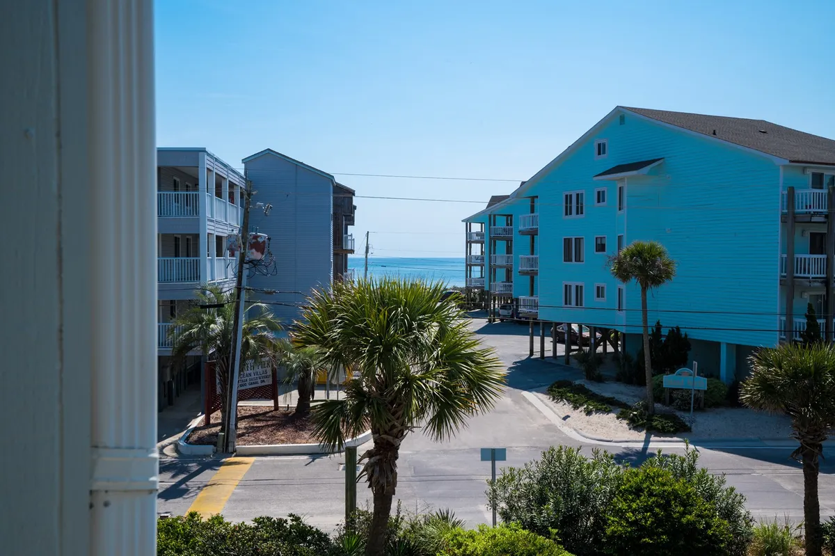 This image captures a sweeping view of the sea with palm trees dotting the scene and charming beachside buildings in the background. The setting includes people strolling along the sandy shore, enjoying beachfront dining, and exploring the nearby shops. The overall mood is inviting, evoking the quintessential beach experience at Carolina Beach.