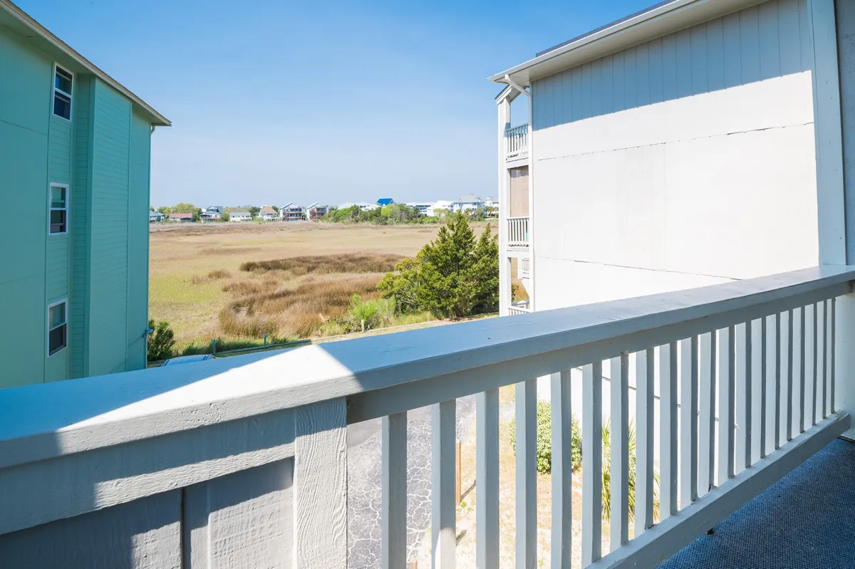 A private balcony with a clear railing reveals expansive marshland views and distant houses. The peaceful setting invites quiet moments of reflection or enjoying a beverage while soaking up the scenery.
