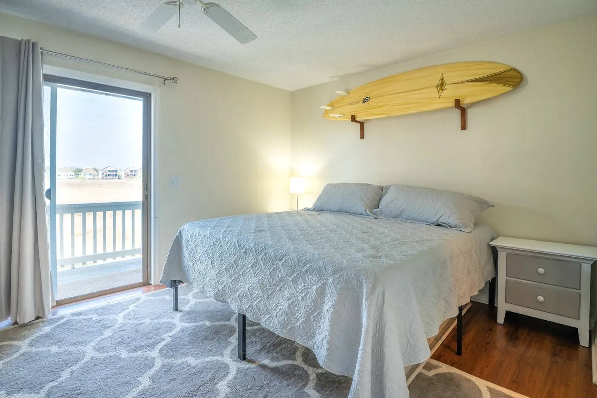 A tranquil bedroom featuring a cozy bed positioned near a large window, with a surfboard as a decorative accent. Soft natural light fills the space, creating an inviting atmosphere for rest and relaxation.