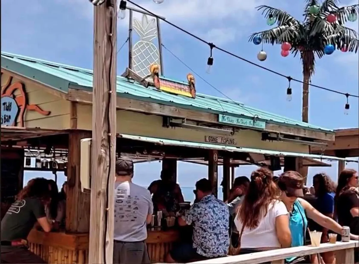 This exterior image highlights a lively tiki bar area adorned with string lights and palm tree decorations. The scene suggests a vibrant community spot where guests can socialize, enjoy drinks, and participate in evening gatherings, all set against a backdrop of tropical decor.