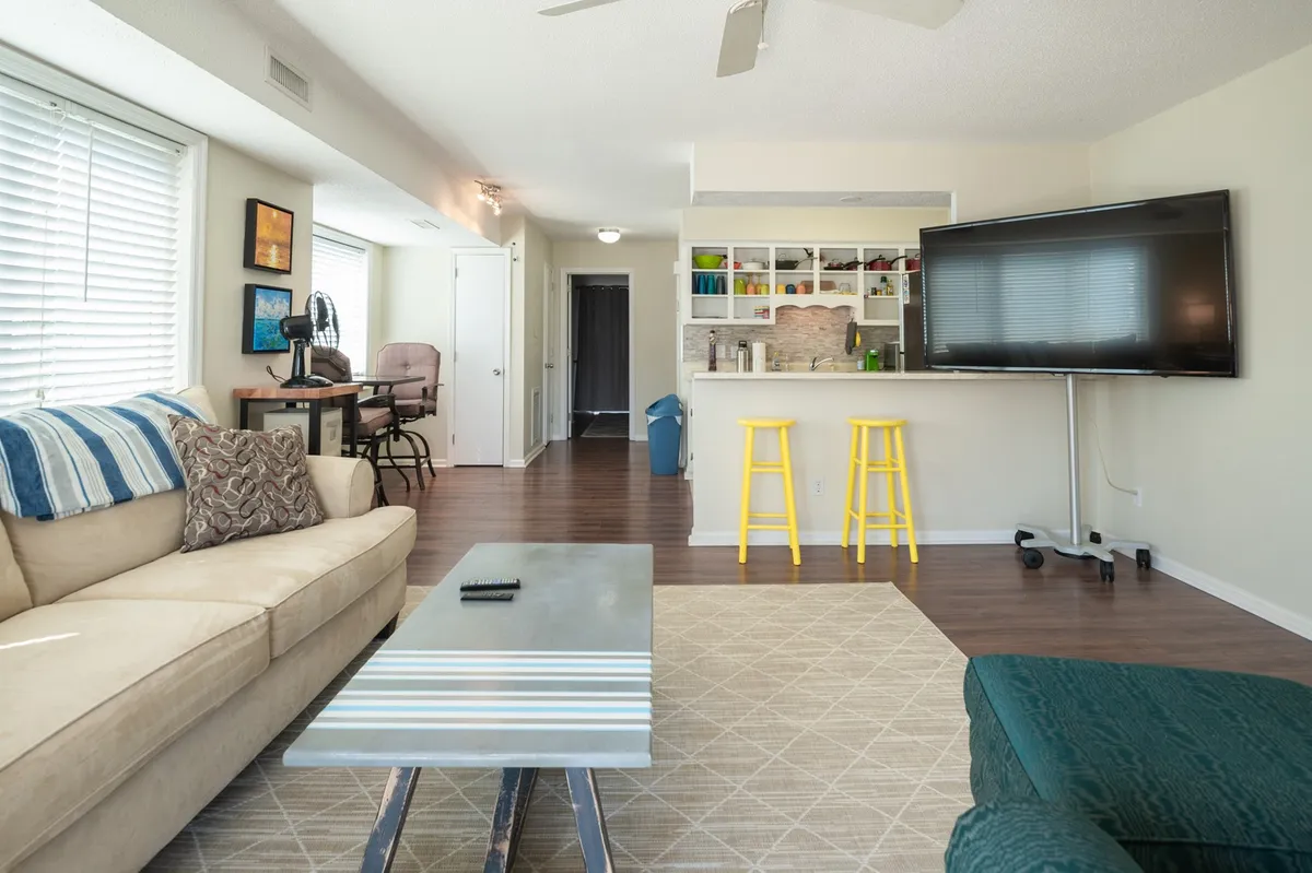 This image captures a vibrant living space with a plush sofa, a rustic coffee table adorned with surf-themed accents, a large TV, bar stools for casual dining, and coastal-inspired artwork. The mood is cozy and adaptable, perfect for both downtime and social gatherings.