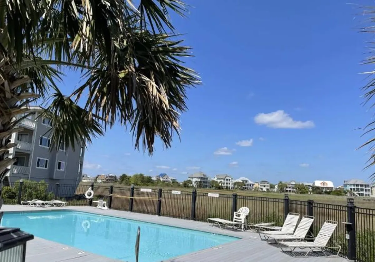 A tranquil community swimming pool surrounded by palm trees and nearby houses. The water glistens in the sunlight, with inviting lounge chairs lining the pool deck and a backdrop of marshland that hints at a peaceful, natural setting.