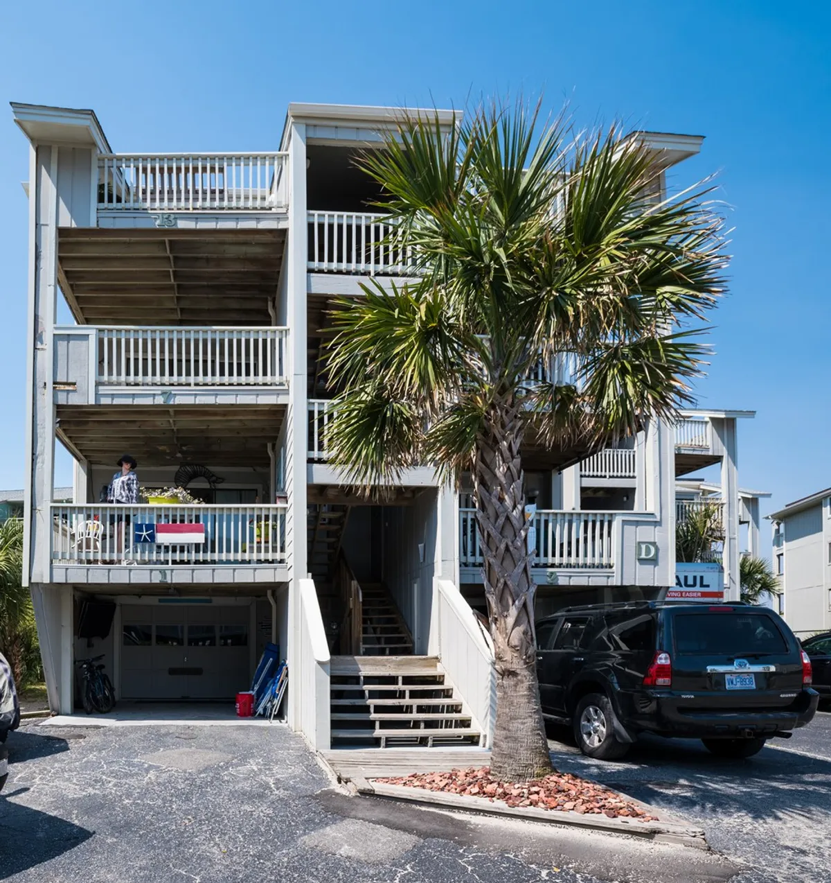 The exterior of a property featuring palm trees, balconies with residents relaxing, and cars parked below. A person enjoys downtime on the balcony as sunset light filters through, setting a relaxed and welcoming mood. The scene hints at the proximity to local attractions and the convenience of stepping right outside into the action.